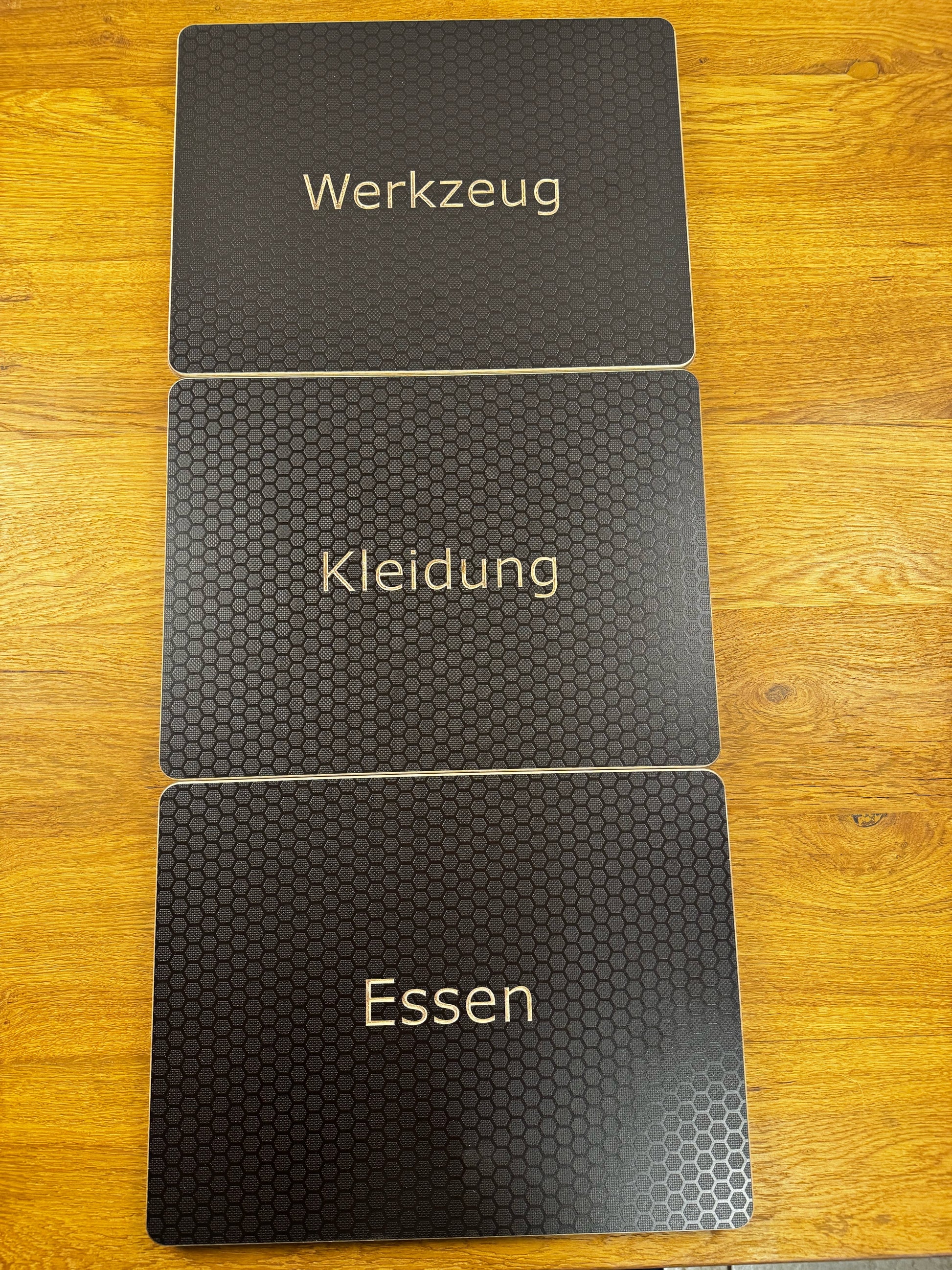 Drei Eurobox-Deckel Hexagon mit eingefräster Beschriftung Werkzeug, Kleidung und Essen, nebeneinander auf Eichendielen